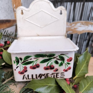 以下が含まれることがあります： 白いヴィンテージのマッチホルダー。チェリーのデザインと黒字で「ALLUMETTES」の文字が描かれています。ホルダーには蓋があり、背面にひし形の装飾があります。緑と赤いベリーに囲まれています。