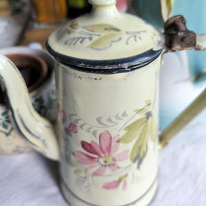 Könnte beinhalten: Cremefarbene Vintage-Emaille-Kaffeekanne mit Deckel und Griff. Die Kanne ist mit rosa und grünen floralen Designs verziert. Der Rand und der Deckel sind mit einem dunkelblauen Rand versehen. Die Kanne hat einen Ausguss und einen Griff.