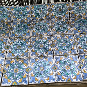 Op de afbeelding: Een verzameling vierkante keramische tegels met een bloemmotief. De tegels hebben een herhalend patroon van blauwe, turquoise en gele bloemen op een witte achtergrond. De tegels zijn gerangschikt op een houten oppervlak.