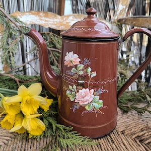 Könnte beinhalten: Eine braune Vintage-Kaffeekanne aus Emaille mit floralem Design. Die Kanne hat einen Deckel, einen Ausguss und einen Griff. Das florale Design umfasst rosa und weiße Rosen, grüne Blätter und blaue Akzente. Gelbe Narzissen und Grünzeug befinden sich im Vordergrund.