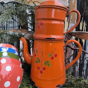 Può includere: Una caffettiera arancione in smalto con coperchio, beccuccio e manico. La caffettiera è decorata con un motivo a ramo di ciliegio. Una pila di ciotole rosse a pois bianchi è visibile in primo piano.