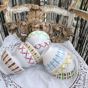 Peut inclure: Ensemble de bols en céramique blanche ornés de motifs géométriques colorés. Les bols présentent des motifs rouges, jaunes, verts et bleus. Ils sont disposés sur une nappe en dentelle blanche, évoquant un style vintage ou artisanal.