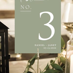May include: Sage green table number card with the number 3 in white. The card reads "table NO." in script and block letters. Below the number is the text "DANIEL - JANET 07.13.20xx". The card is held by a gold clip.