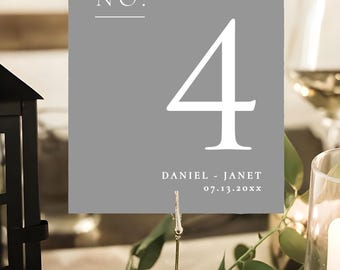 Può includere: Un segnaposto grigio con il numero 4 bianco. La carta presenta le parole "table NO." in alto e i nomi "DANIEL - JANET" e una data in basso. La carta è tenuta da una clip dorata.