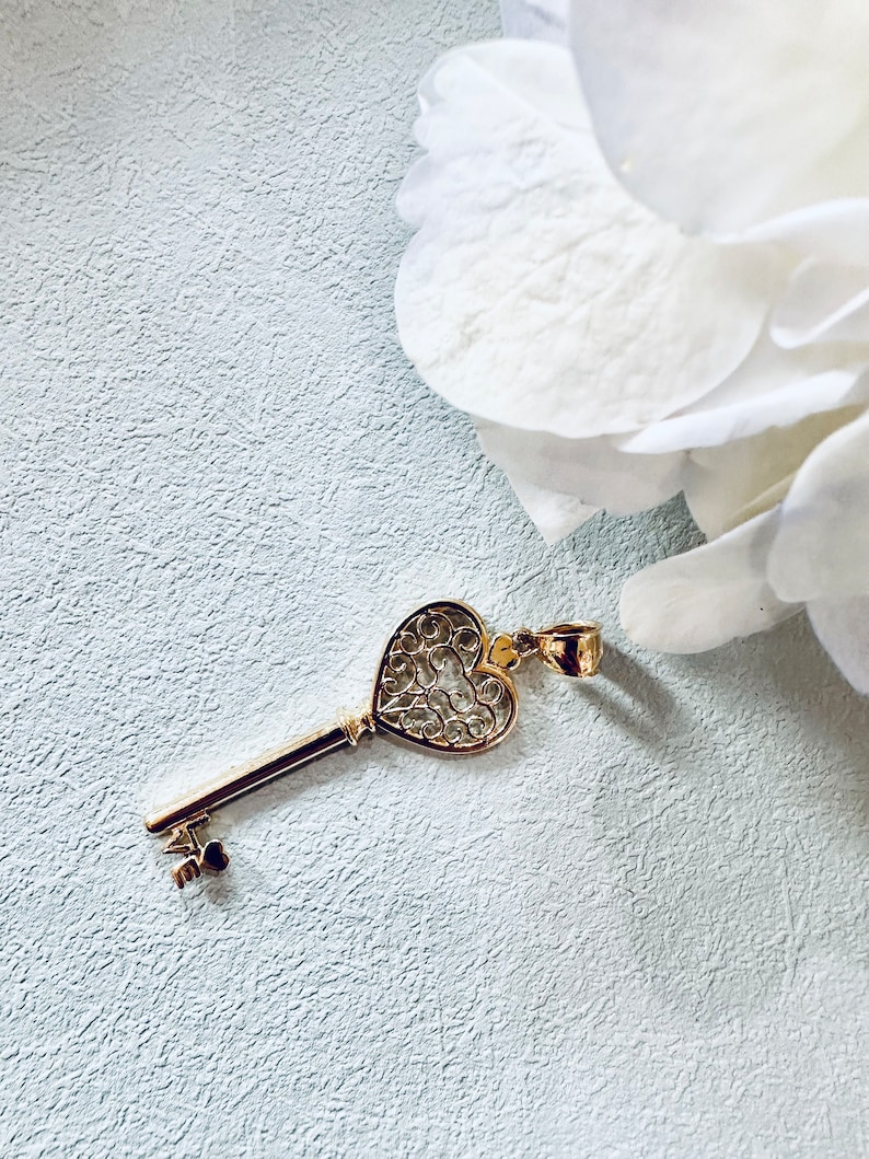 Op de afbeelding: Een gouden hartvormige sleutel hanger met ingewikkelde filigraan details. De sleutel heeft een lange, dunne steel en een kleine, ronde knop aan het einde.