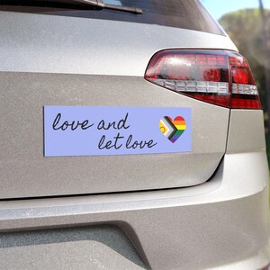Pode incluir: Um autocolante retangular azul-claro para para-choques com o texto em cursivo preto love and let love seguido de um ícone de coração com as cores da bandeira Progress Pride, incluindo faixas vermelhas, laranjas, amarelas, verdes, azuis, violetas, castanhas, pretas, brancas, rosas e azul-claras.