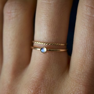 May include: Two gold rings are shown on a finger. The bottom ring has a small, round, light blue gemstone. The top ring has a twisted rope design. The rings are delicate and minimalist.