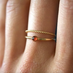 May include: Two gold rings on a finger. The top ring is a twisted band. The bottom ring has a small, round, orange gemstone.