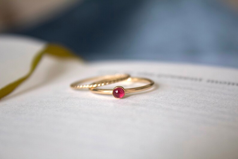 May include: A gold ring with a small, round, pink gemstone set in the center. The ring is on a white surface.