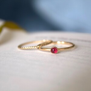 May include: Two gold rings, one with a small, round, pink gemstone. The rings are on a white surface.