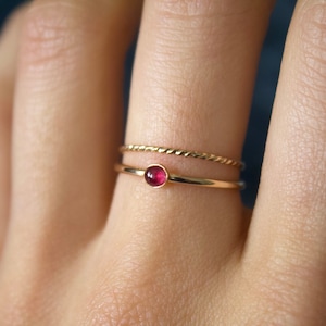 May include: A gold ring with a small, round, red gemstone. The ring is worn on a finger with a second, thinner gold band.
