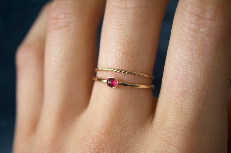 May include: A gold ring with a small, round, red gemstone. The ring is worn on a finger with a second, thinner gold band.