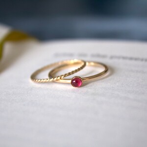 May include: Two gold rings, one with a twisted band and the other with a single red gemstone, are stacked on a white surface.