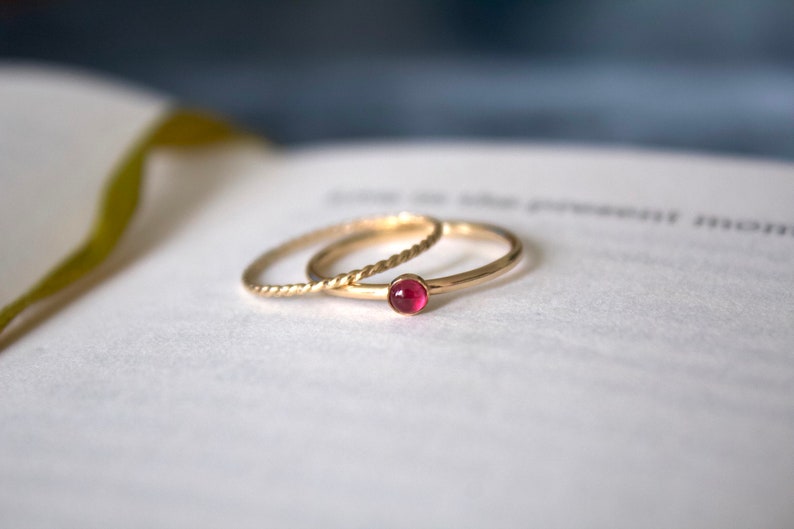 May include: Two gold rings, one with a twisted band and the other with a single red gemstone, are stacked on a white surface.