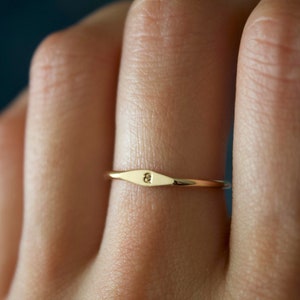 May include: A thin gold band ring with a flat, rectangular top. The top of the ring has a small, engraved design.