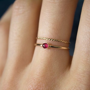 May include: A gold ring with a small, round, red gemstone. The ring is worn on a finger with a thin gold band on the finger next to it.