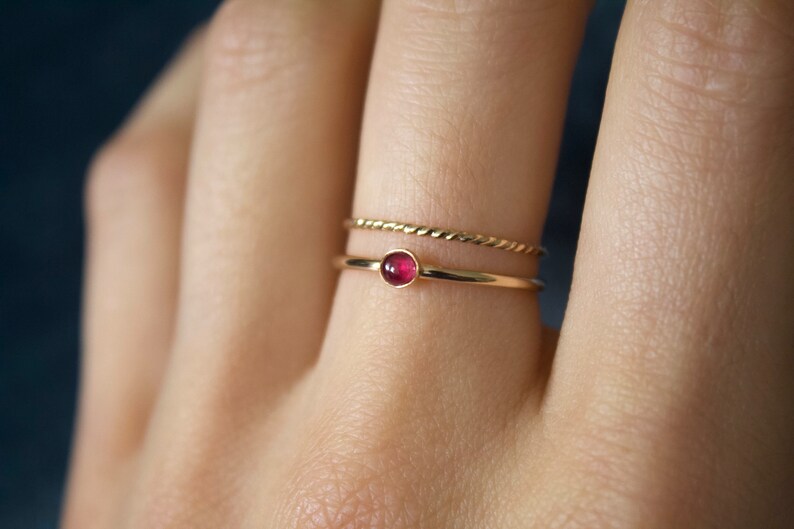 May include: A gold ring with a small, round, red gemstone. The ring is worn on a finger with a thin gold band on the finger next to it.