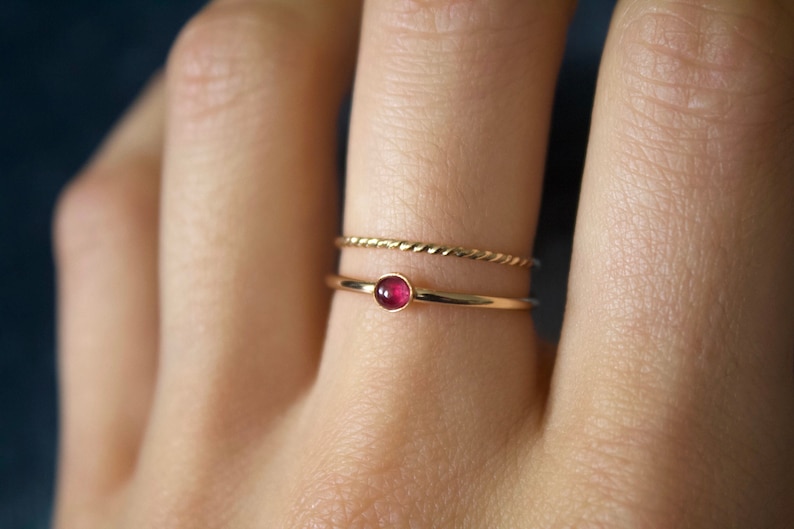 May include: A gold ring with a small, round, red gemstone. The ring is worn on a finger with a second, thinner gold band.