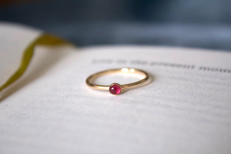 May include: A simple gold band ring with a small, round, pink gemstone set in the center.