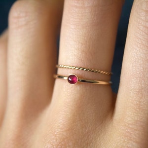 May include: A gold ring with a small, round, red gemstone. The ring is worn on a finger with a thin gold band on the finger next to it.