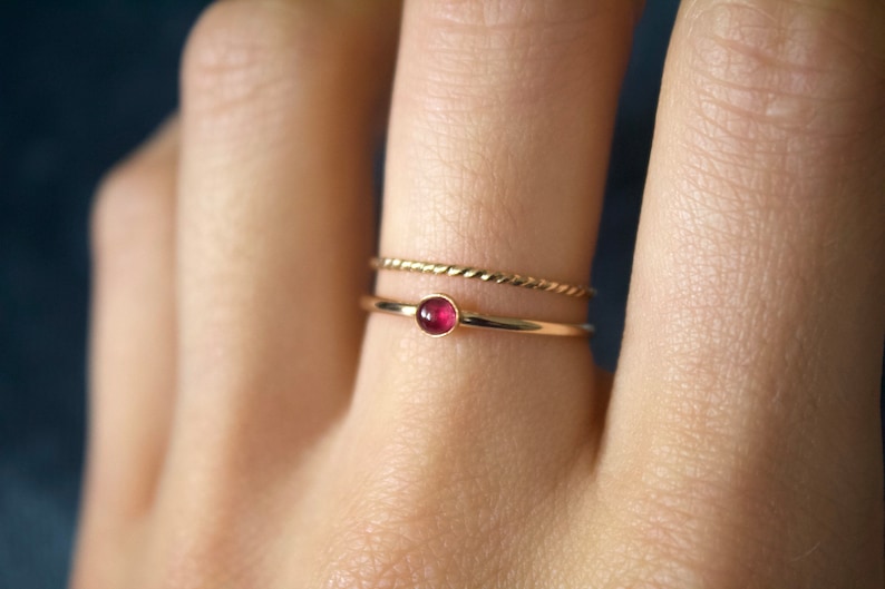 May include: A gold ring with a small, round, red gemstone. The ring is worn on a finger with a thin gold band on the finger next to it.