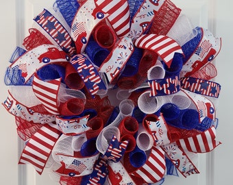 May include: A patriotic wreath with red, white, and blue ribbons. The wreath features striped and star-patterned ribbons, along with ribbons depicting a red truck and fireworks. The wreath is designed for the Fourth of July or other patriotic holidays.