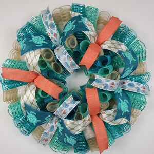 May include: A round decorative wreath featuring turquoise, coral, and tan mesh ribbons. The wreath is adorned with patterned ribbons displaying sea turtles and seashells. Coral-colored bows are evenly spaced around the wreath, creating a coastal theme.
