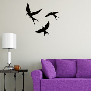 May include: Three black silhouette bird wall decals in flight, mounted on a white wall above a purple sofa with two pillows.