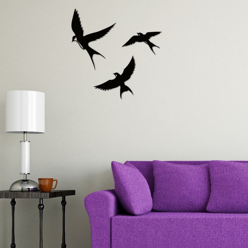 May include: Three black silhouette bird wall decals in flight, mounted on a white wall above a purple sofa with two pillows.