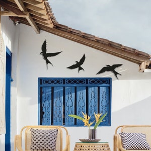May include: Three black metal bird silhouettes mounted on a white wall with a blue window shutter. The birds are in flight and are arranged in a staggered pattern. The wall is painted white and the window shutter is painted blue.