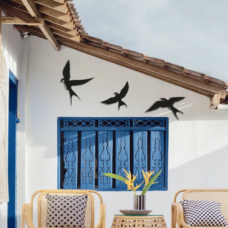 May include: Three black metal bird silhouettes mounted on a white wall with a blue window shutter. The birds are in flight and are arranged in a staggered pattern. The wall is painted white and the window shutter is painted blue.