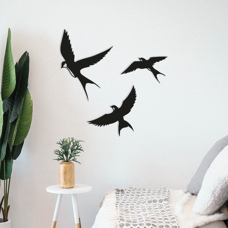May include: Three black metal silhouette wall decor pieces in the shape of birds. The birds are arranged in a staggered pattern on a white wall. The birds are all facing the same direction.