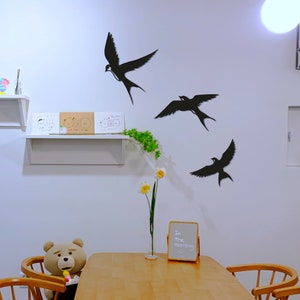 May include: A white wall with three black swallow silhouettes, a white shelf with three drawings, a vase with yellow flowers, a brown teddy bear sitting on a wooden chair, and a small black chalkboard with the text "In the morning" on a wooden stand.