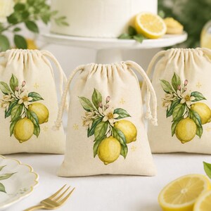 May include: Three off-white fabric drawstring pouches featuring a printed botanical illustration of yellow lemons, green leaves, and white blossoms. Each pouch has a matching cream-colored cord closure at the top, centered on a clean surface.