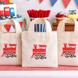 Puede incluir: Tres bolsas de mano de lona color crema con gráficos de trenes rojos. Las bolsas están sobre una superficie a rayas rojas y blancas, con una tarta y cupcakes con temática de trenes en el fondo. Decoraciones de fiesta con banderines y un recipiente de palomitas de maíz a rayas rojas y blancas.