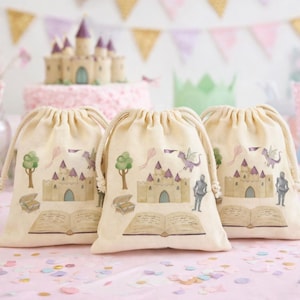 May include: Three beige drawstring bags with a castle design, perfect for a party. The bags feature a castle, dragon, knight, and open book illustration. A cake and cupcakes are in the background, with confetti on the table.