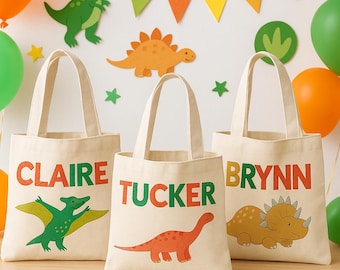 Puede incluir: Tres bolsas de mano de lona beige con nombres en letras naranjas y verdes. Cada bolsa presenta una ilustración de dinosaurio diferente en verde y naranja. Globos y decoraciones de dinosaurios en el fondo.