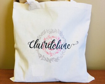Puede incluir: Bolso tote de color crema con el texto "Clairdelune" en fuente cursiva, rodeado por un diseño de corona con hojas rosas y grises. El bolso tiene dos asas y está hecho de una tela resistente.