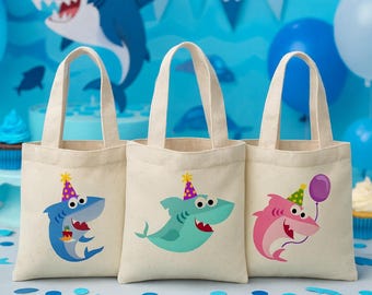 May include: Three beige canvas tote bags with shark illustrations. Each shark wears a party hat and has a different color scheme: blue, teal, and pink. The background features blue balloons and a banner that says "PARTY".