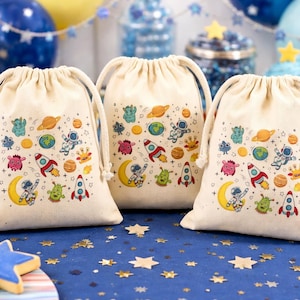 May include: Three cream-coloured drawstring bags with a colourful space-themed design featuring rockets, planets, astronauts, and aliens. The bags are on a blue surface with star-shaped confetti, with cupcakes and balloons in the background.