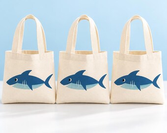 May include: Three off-white canvas tote bags arranged in a row, each featuring a centered graphic of a blue shark with a light blue underbelly. The bags have two matching fabric handles and a square base, presented against a light blue background.