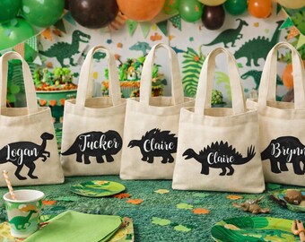 Puede incluir: Cinco bolsas de mano de lona beige con siluetas de dinosaurios negros y nombres. Las bolsas están sobre una superficie verde con decoraciones de fiesta con temática de dinosaurios, incluidos globos y platos. Ideales para una fiesta de cumpleaños con temática de dinosaurios.