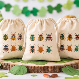 Puede incluir: Tres bolsas beige con cordón y ilustraciones de insectos coloridos. Las bolsas están dispuestas sobre una rodaja de madera, con hojas de papel verdes y elementos decorativos en el fondo. Ideales para regalos.