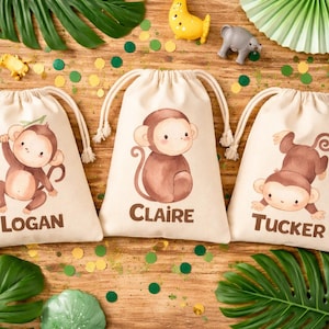 May include: Three beige drawstring bags with cartoon monkey illustrations and names: Logan, Claire, and Tucker. The bags are on a wooden surface with jungle-themed decorations, including toy animals and green confetti.