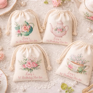 May include: Four cream-colored drawstring bags with floral designs and the text "Lucy's 1st Birthday Par-tea." The bags feature illustrations of teapots and teacups with pink and green floral accents. The scene includes teacups, tea, and macarons.