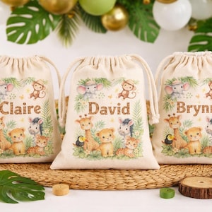 May include: Three beige drawstring bags with jungle animal illustrations and names: Claire, David, and Brynn. Each bag features a giraffe, lion, zebra, and other animals. The bags are on a woven mat with decorative items.