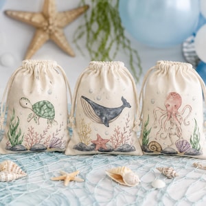 Könnte beinhalten: Drei cremefarbene Stoffbeutel mit Kordelzug, die jeweils mit einer Aquarellillustration eines Meerestieres versehen sind. Die linke Tasche zeigt eine grüne Meeresschildkröte, die mittlere einen Blauwal und die rechte einen rosafarbenen Oktopus.