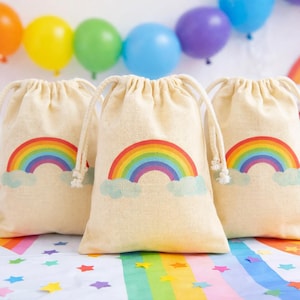 Könnte beinhalten: Drei beigefarbene Kordelzugbeutel mit Regenbogen-Design und blauen Wolken. Die Beutel stehen auf einem Tisch im Regenbogen-Thema, mit Luftballons und Partyzubehör im Hintergrund. Ideal für Partygeschenke.