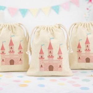 Könnte beinhalten: Drei beige Kordelzugbeutel mit einem rosa Schlossdesign. Jeder Beutel zeigt ein Schloss mit rosa Türmen, blauen Fahnen und einer braunen Tür. Die Beutel liegen auf einer weißen Oberfläche mit Konfetti und Partyzubehör.