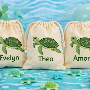 Peut inclure: Trois sacs beiges à cordon de serrage avec des illustrations de tortues vertes et des noms : Evelyn, Théo et Amora. Les sacs sont posés sur une surface bleue avec des décorations sur le thème des tortues.